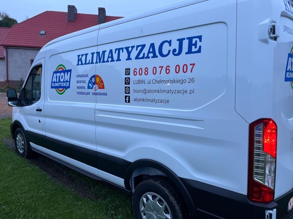 Biały samochód dostawczy z dużym napisem 'KLIMATYZACJE' i danymi kontaktowymi firmy Atom Klimatyzacje, zaparkowany przed budynkiem.