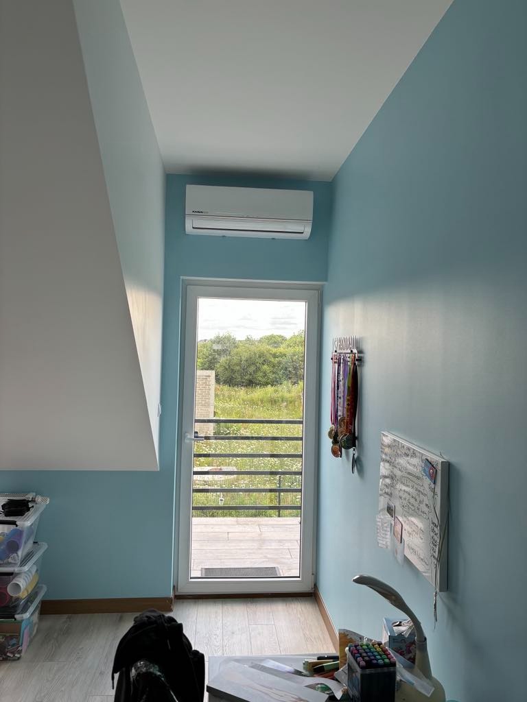 Błękitny pokój na poddaszu z zamontowaną klimatyzacją Kawai nad drzwiami balkonowymi z widokiem na zieleń, obok wiszą medale i tablica suchościeralna.