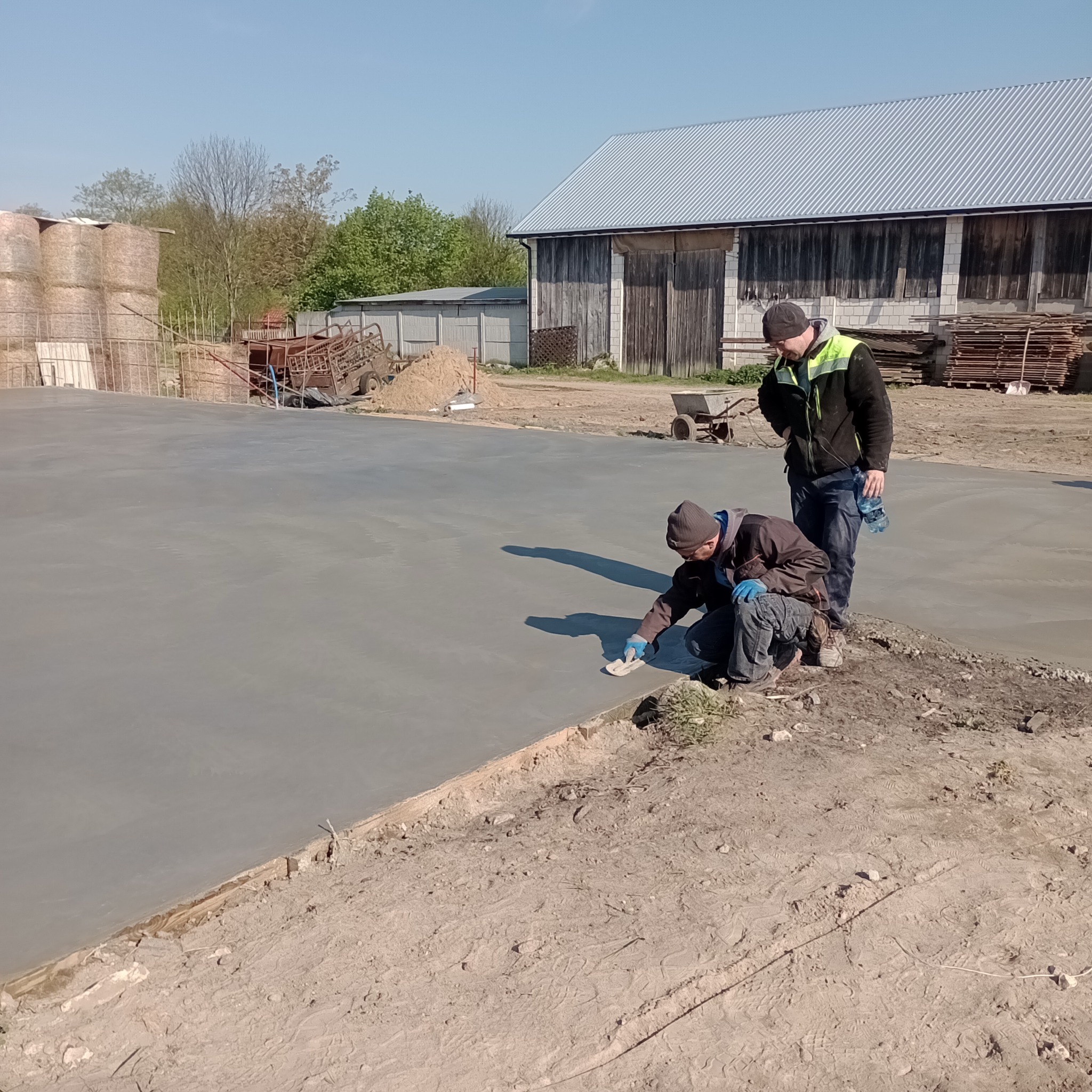 Dwóch pracowników wygładza krawędzie świeżo wylanej betonowej posadzki na zewnątrz budynku gospodarczego z belami siana w tle.
