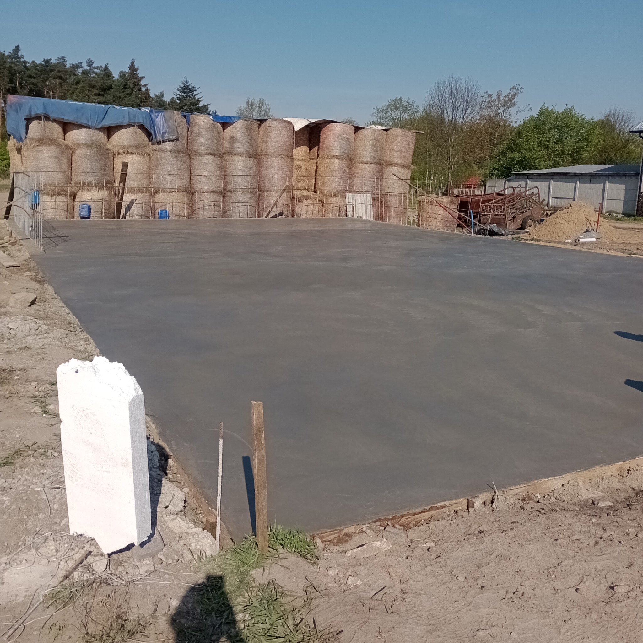 Wylana, świeża, gładka betonowa posadzka na zewnątrz, obok stogi siana zabezpieczone plandeką, widoczne szalunki.