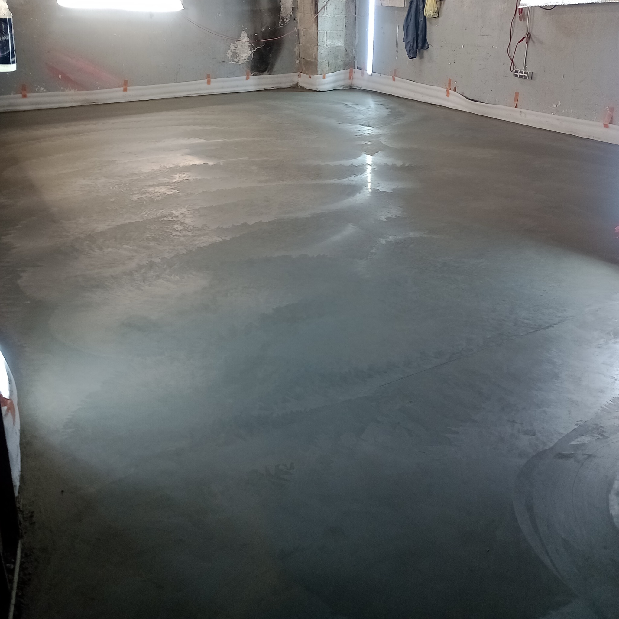 Świeżo wylana, gładka, szara posadzka w pomieszczeniu z widocznymi narożnikami zabezpieczonymi pianką i elementami instalacji na ścianach.