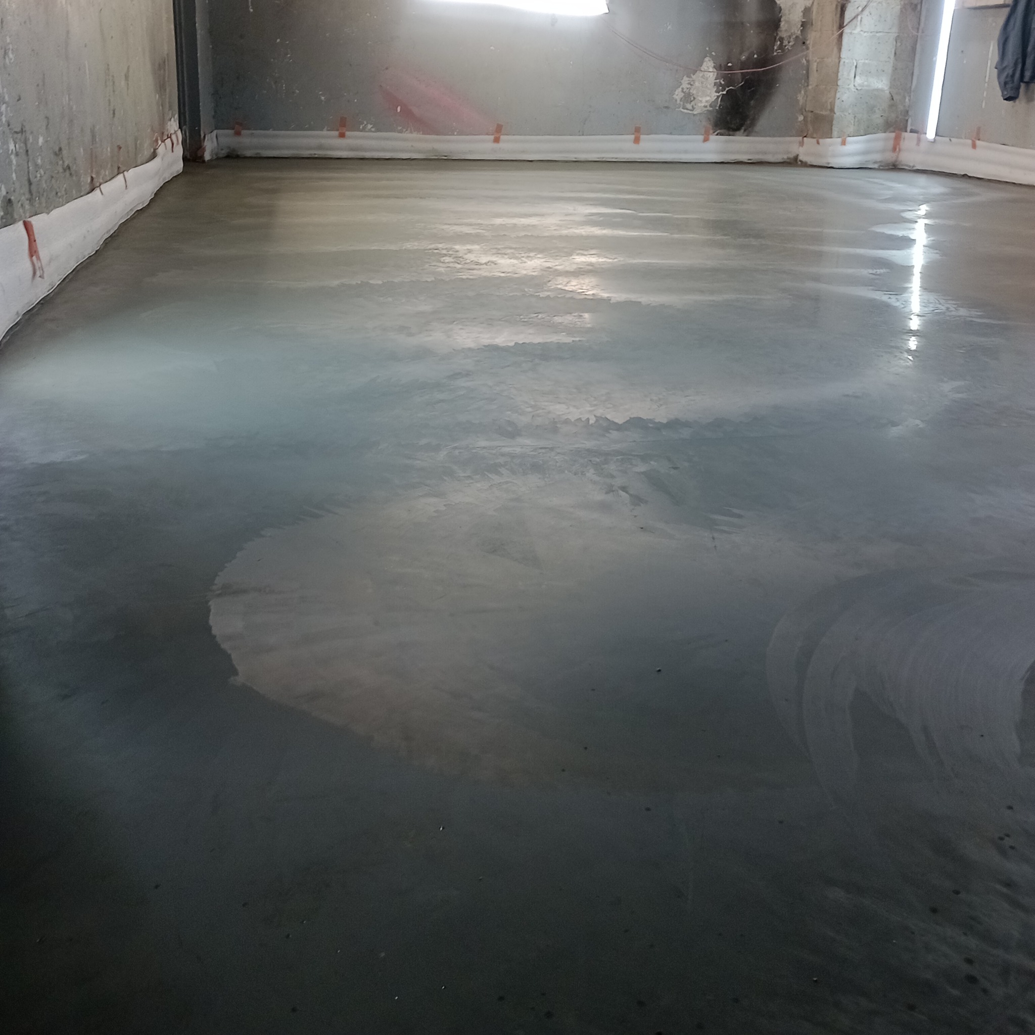 Świeżo wylana, mokra, szara posadzka w przemysłowym pomieszczeniu, zabezpieczona białą folią przy ścianach, widoczne ślady po wygładzaniu.