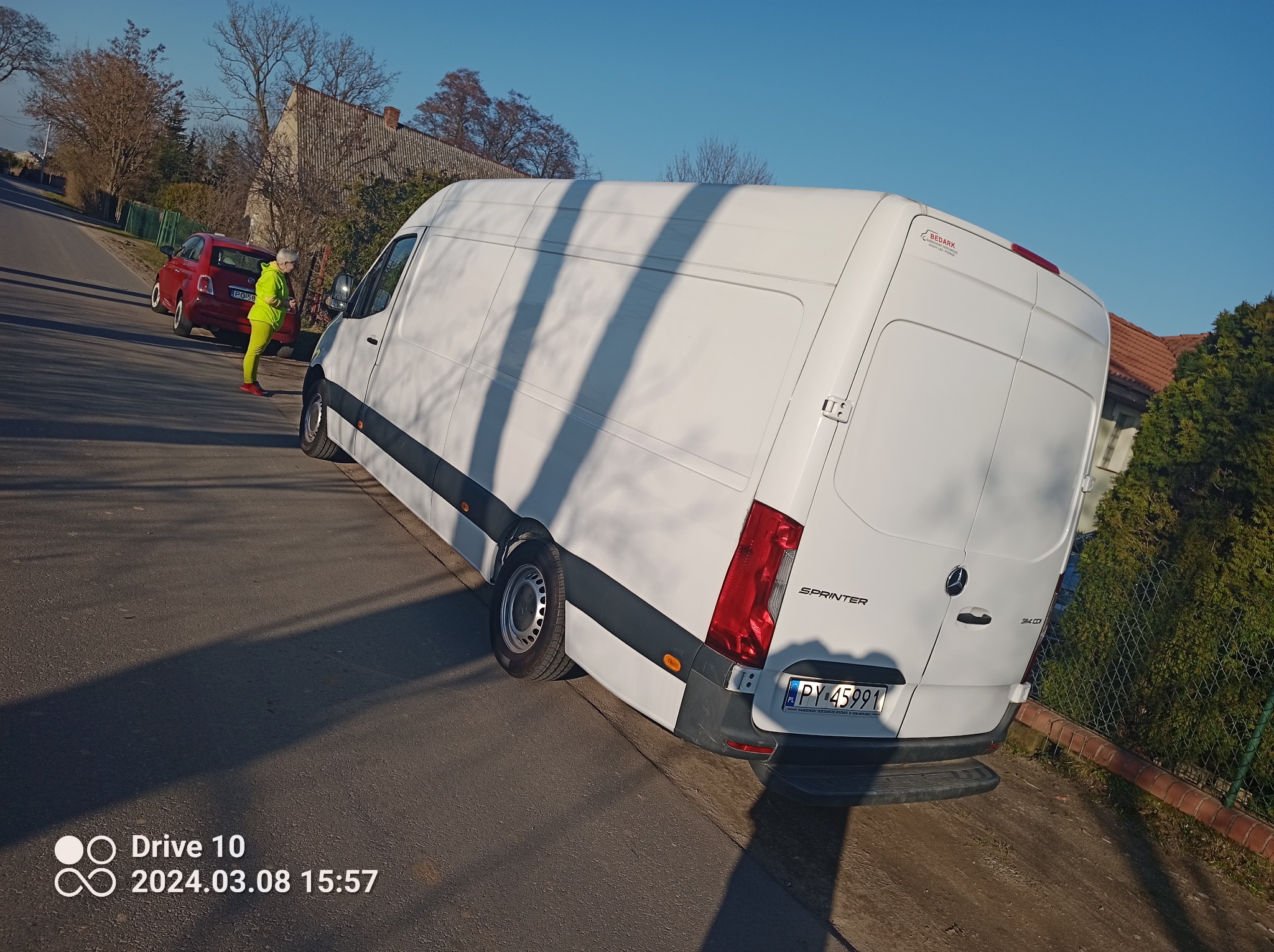 Biały samochód dostawczy marki Mercedes-Benz Sprinter zaparkowany na pochyłej drodze, z widoczną osobą w jaskrawożółtym kombinezonie stojącą obok pojazdu i czerwonym samochodem osobowym w tle.
