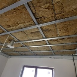 Slawbud - Sufit podwieszany w trakcie montażu: metalowa konstrukcja z izolacją z wełny mineralnej, widoczne okno i fragment ściany. Ujęcie z dołu, perspektywa.