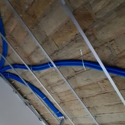 Slawbud - Niebieskie rury przymocowane do metalowej konstrukcji pod sufitem z izolacją, widok z dołu. Elementy instalacji elektrycznej przed zabudową.