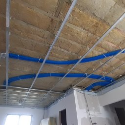 Slawbud - Sufit podwieszany z izolacją i niebieskimi rurami instalacji, widok z dołu. Metalowy stelaż sufitu z elementami oświetlenia w trakcie montażu.