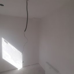 Slawbud - Wnętrze pokoju po malowaniu na biało, widoczne gładkie ściany, okno z promieniami słońca, wystający kabel z sufitu i grzejnik. Minimalizm, surowy stan.