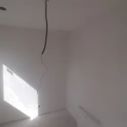 Wnętrze pokoju po malowaniu na biało, widoczne gładkie ściany, okno z promieniami słońca, wystający kabel z sufitu i grzejnik. Minimalizm, surowy stan.