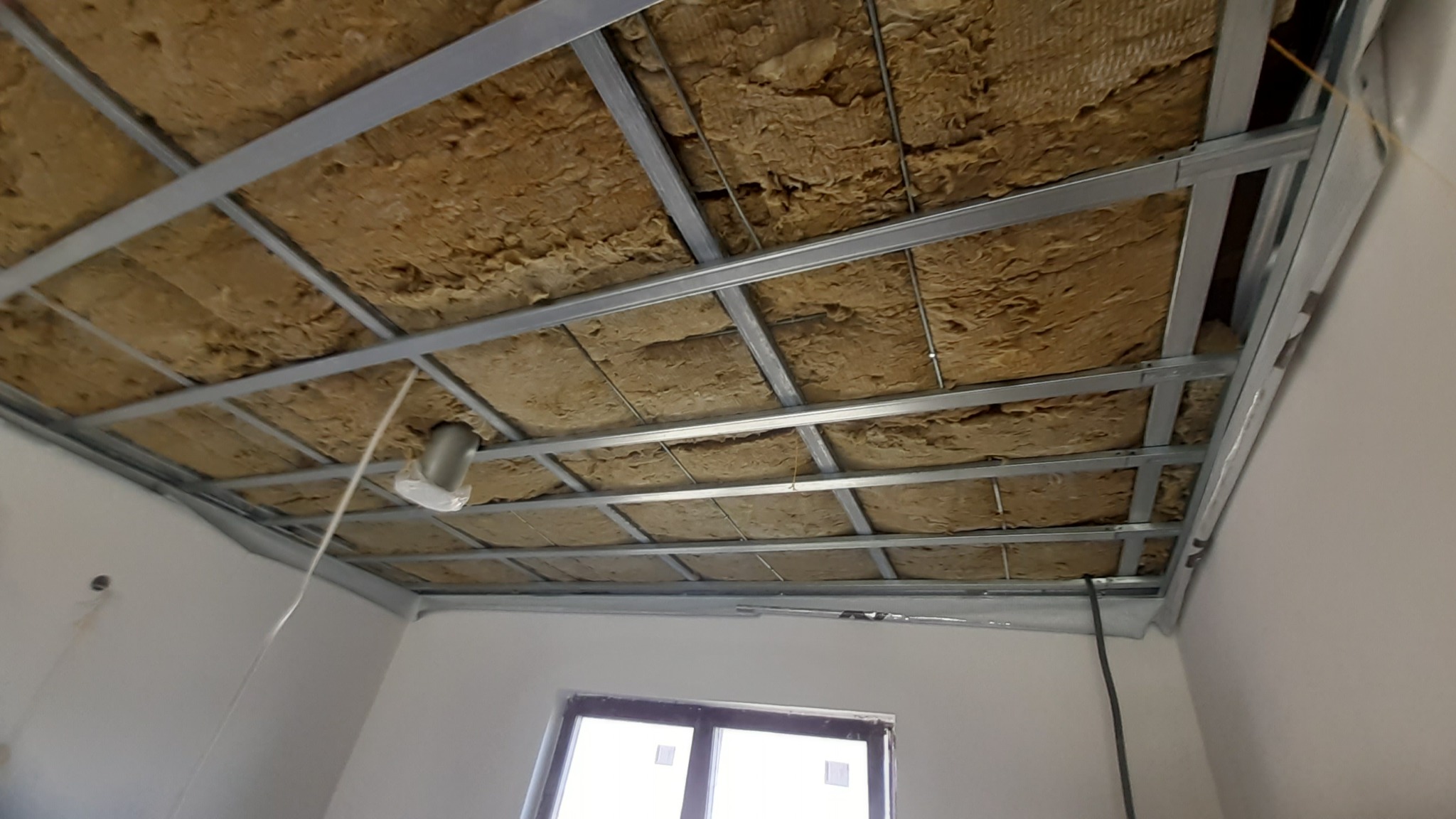 Sufit podwieszany w trakcie montażu: metalowa konstrukcja z izolacją z wełny mineralnej, widoczne okno i fragment ściany. Ujęcie z dołu, perspektywa.