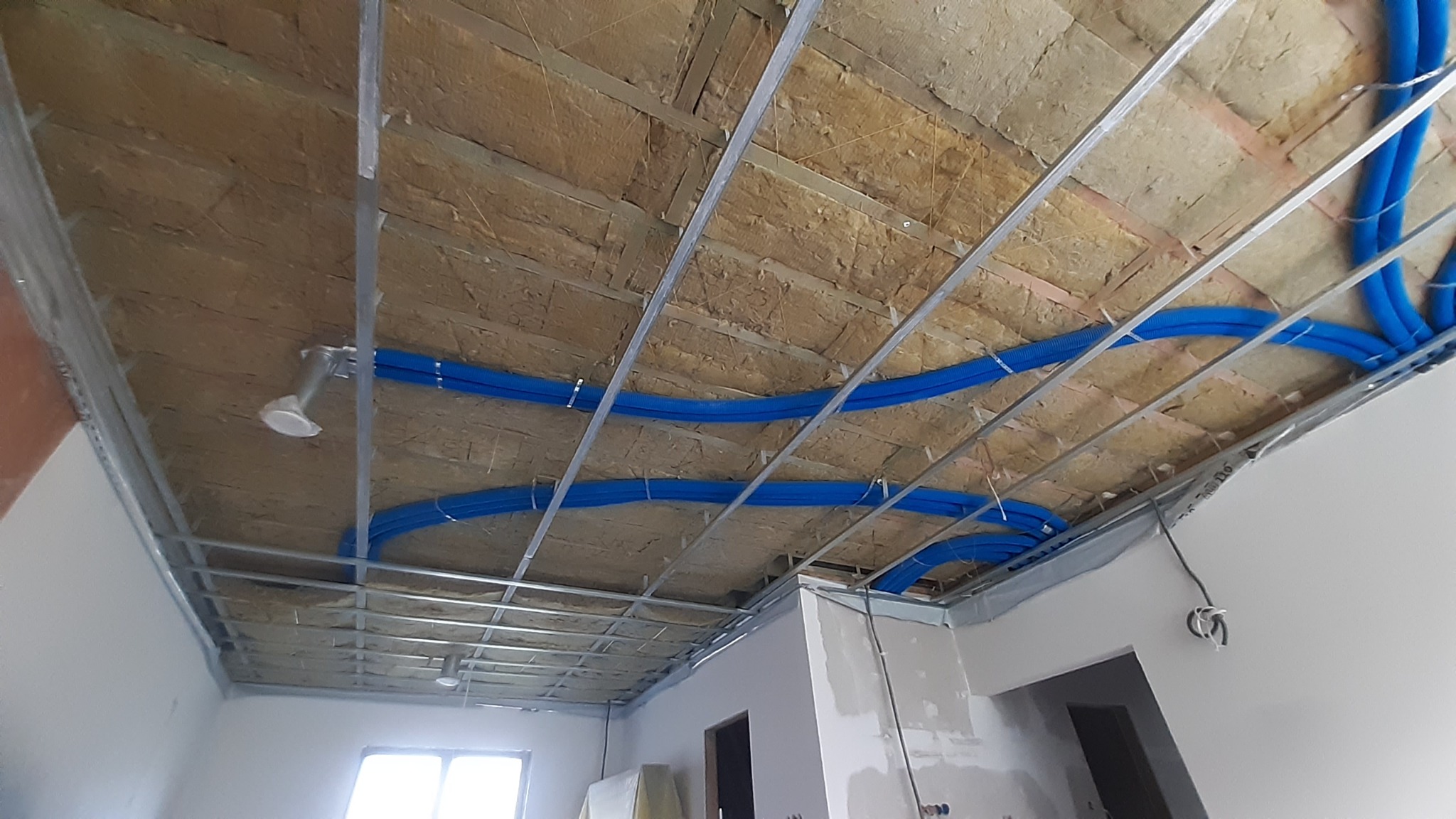 Sufit podwieszany z izolacją i niebieskimi rurami instalacji, widok z dołu. Metalowy stelaż sufitu z elementami oświetlenia w trakcie montażu.