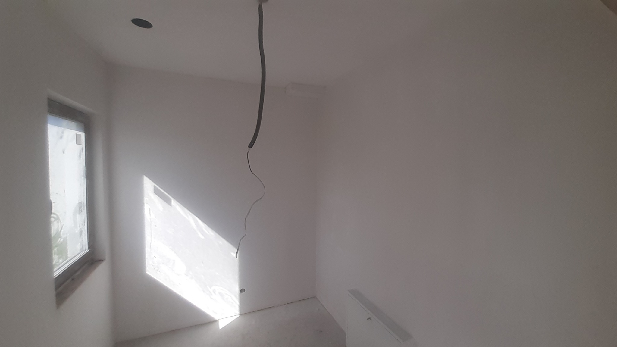 Wnętrze pokoju po malowaniu na biało, widoczne gładkie ściany, okno z promieniami słońca, wystający kabel z sufitu i grzejnik. Minimalizm, surowy stan.