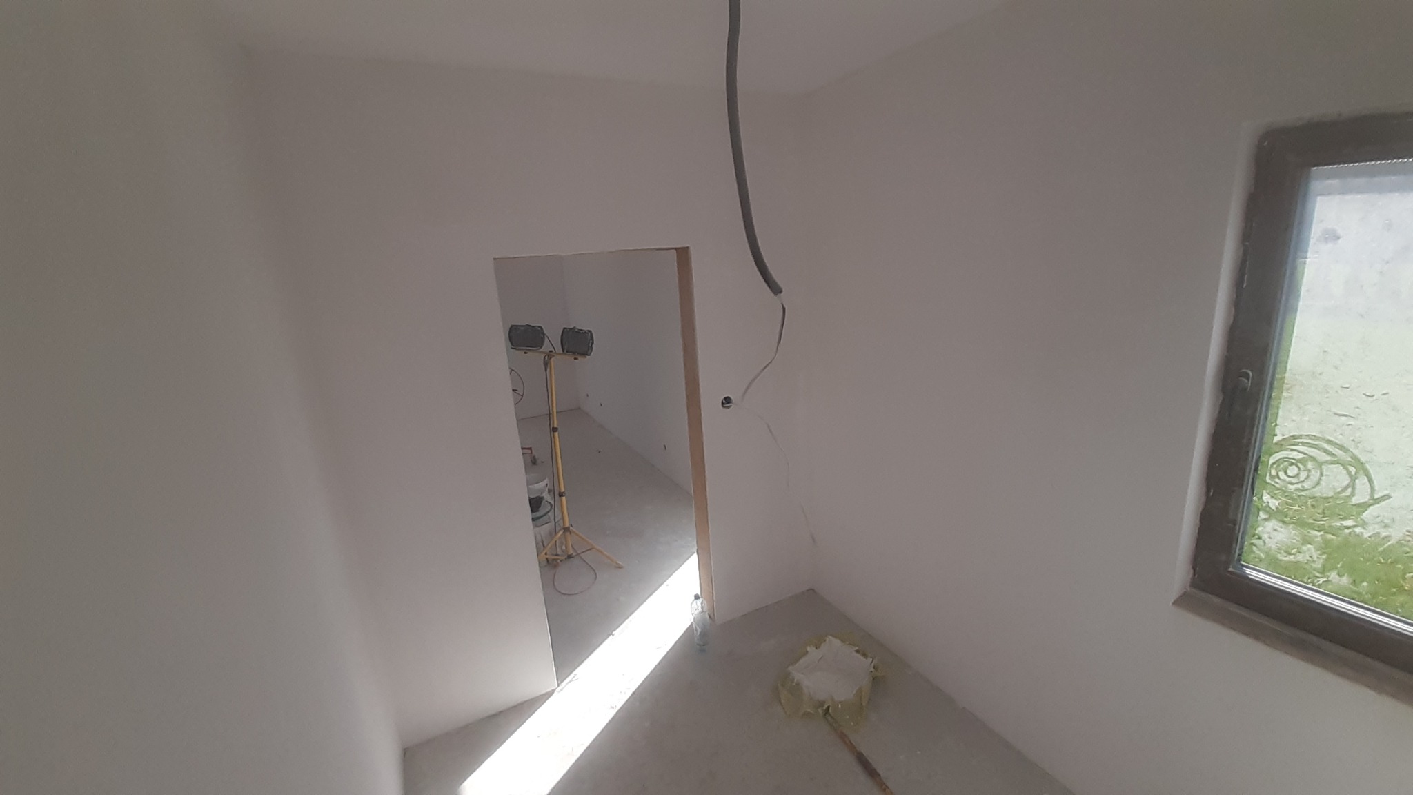 Wnętrze po szpachlowaniu ścian, widoczna lampa budowlana w przejściu, kabel zwisający ze ściany i okno z widokiem na zieleń. Ściany przygotowane do malowania.
