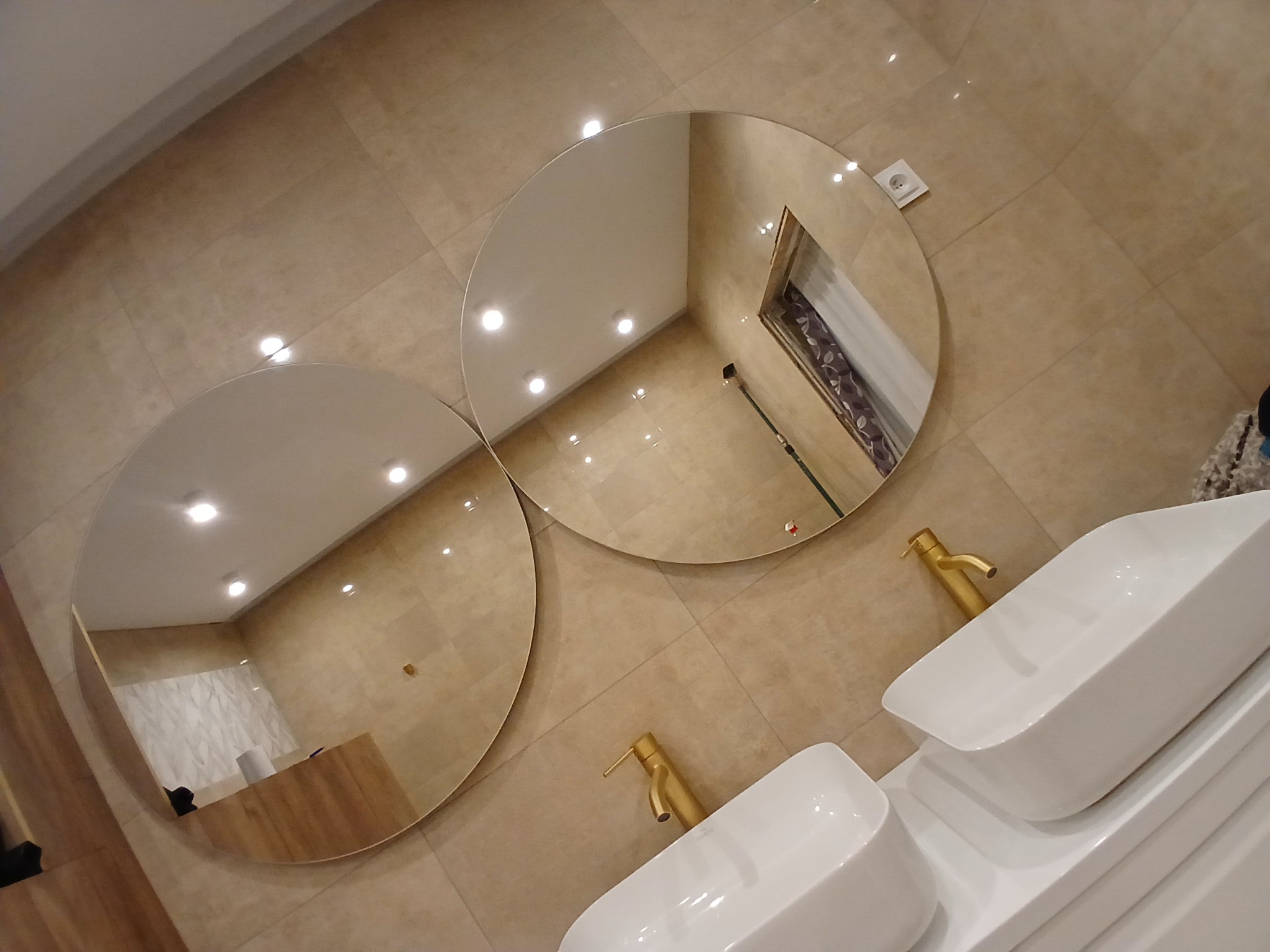 Beżowa łazienka z dwoma okrągłymi lustrami i białymi umywalkami z złotymi bateriami. Widoczne płytki na ścianach i odbicie wnętrza w lustrach.