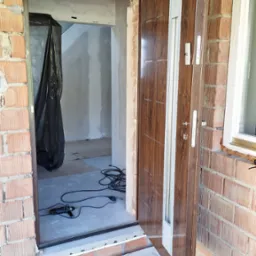 Brązowe drzwi wejściowe z widocznymi elementami montażowymi i narzędziami w surowym stanie wykończenia, ceglana ściana, tymczasowy próg wejściowy, wnętrze w trakcie remontu zabezpieczone folią.