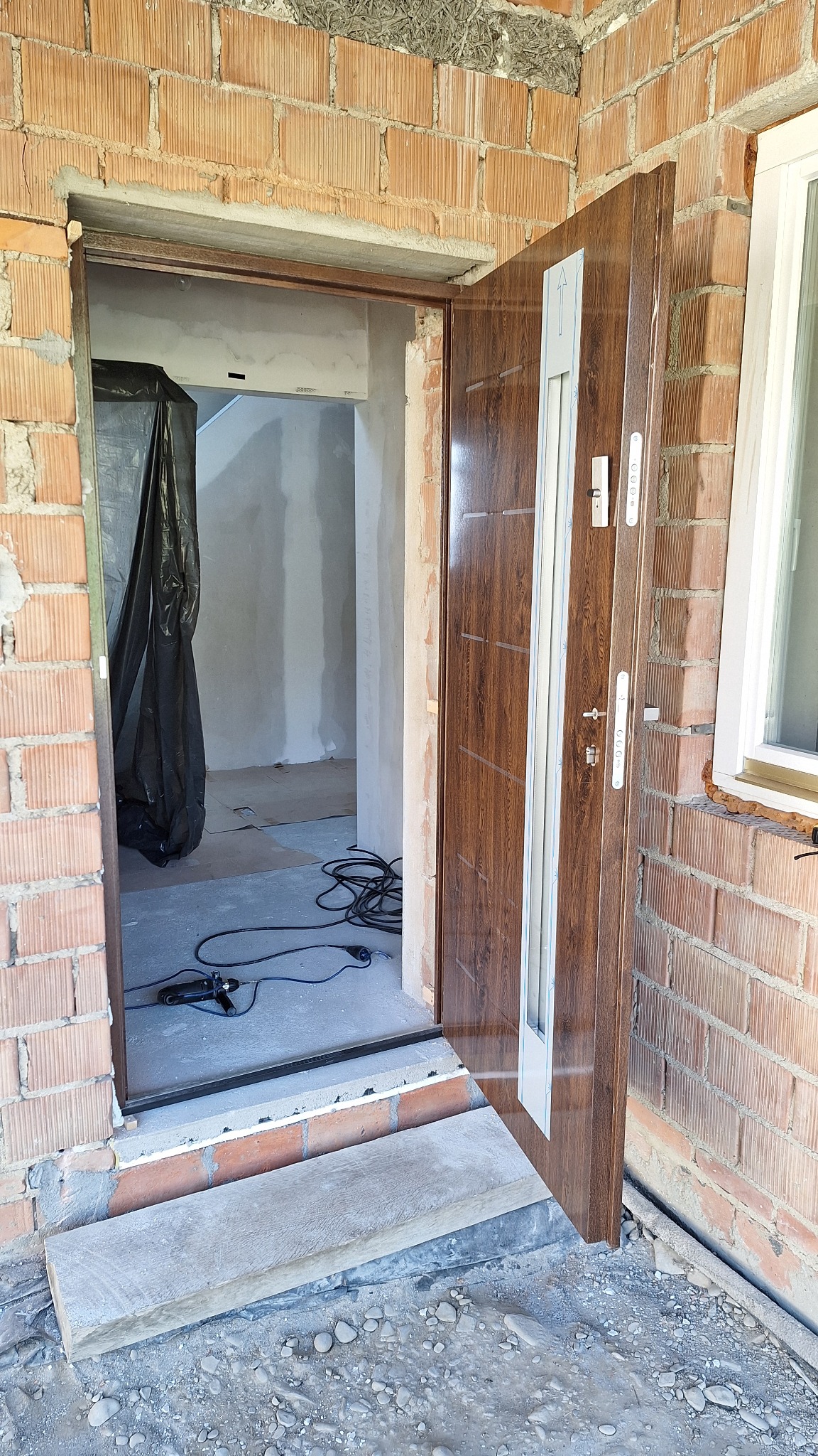 Brązowe drzwi wejściowe z widocznymi elementami montażowymi i narzędziami w surowym stanie wykończenia, ceglana ściana, tymczasowy próg wejściowy, wnętrze w trakcie remontu zabezpieczone folią.