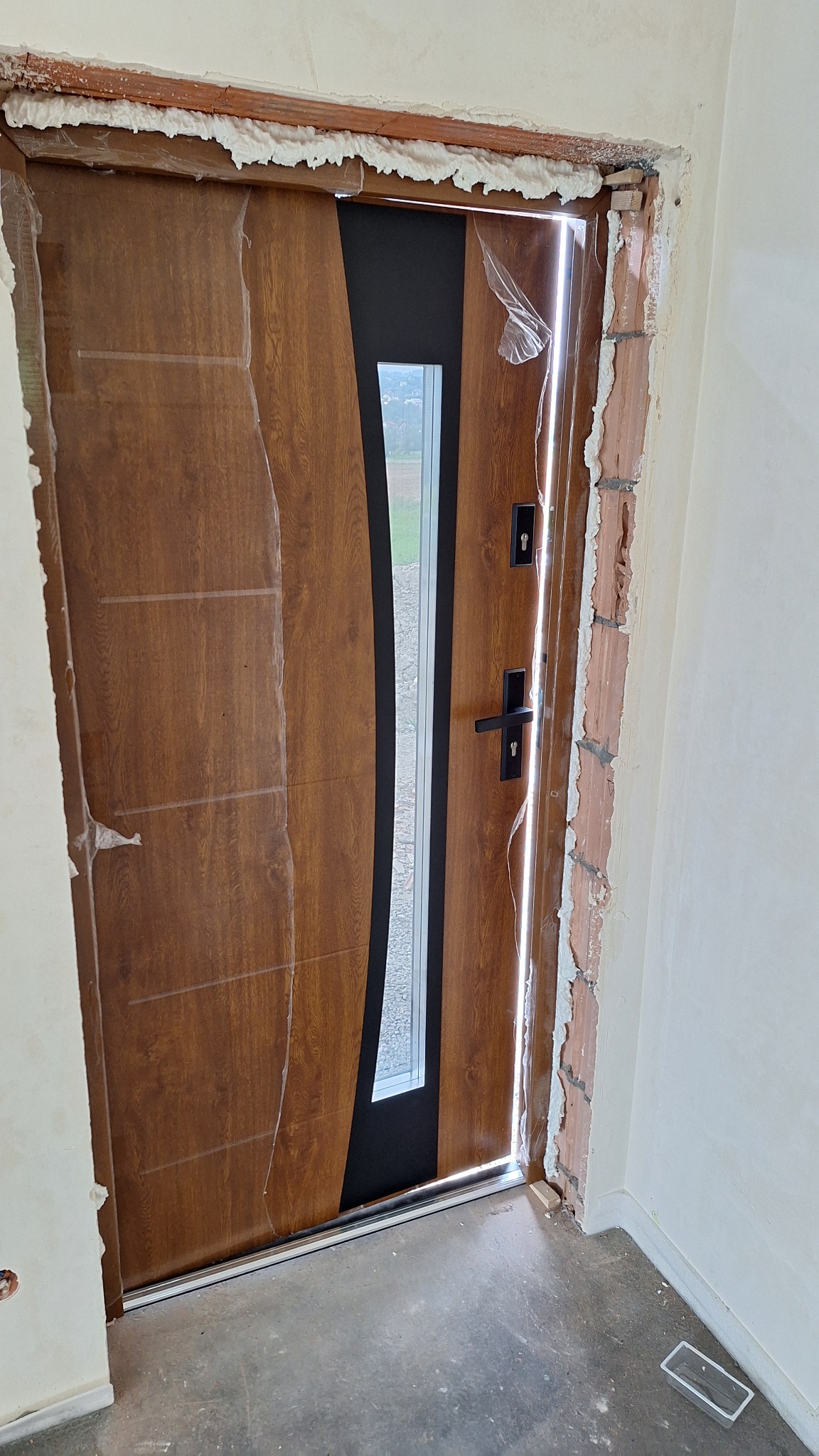 Nowo zamontowane, brązowe drzwi wejściowe z przeszkleniem, zabezpieczone folią ochronną, widoczne pianka montażowa i niedokończone wykończenie ościeżnicy.