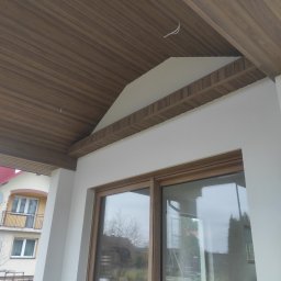 Budowlana - Drewniana podbitka dachowa z widocznymi elementami wykończeniowymi i oświetleniem, fragment elewacji budynku z oknami. Widok z dołu, jasne kolory.