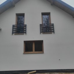 Budowlana - Biała elewacja domu jednorodzinnego z trzema oknami, dwa z czarnymi balustradami. U dołu widoczny fragment fundamentu i ziemi.