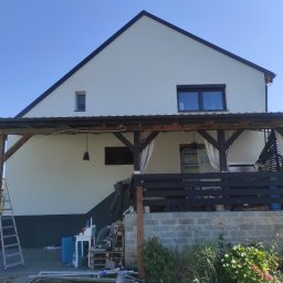 Budowlana - Biały dom z ciemnym dachem i drewnianą werandą. Widoczne rusztowania i materiały budowlane, sugerujące prace wykończeniowe elewacji. Błękitne niebo w tle.