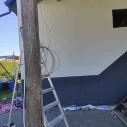 Budowlana - Dwukolorowa elewacja domu w trakcie malowania, widoczna drabina, narzędzia malarskie i zabezpieczenia. Słup drewniany na pierwszym planie.