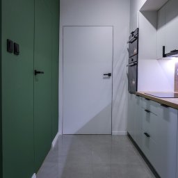 JAVA Architekci - Nowoczesna kuchnia z zabudową w odcieniach szarości i zieleni, minimalistyczne fronty, ukryte drzwi, szara podłoga z płytek, oświetlenie punktowe.
