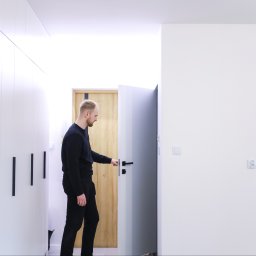 JAVA Architekci - Mężczyzna w czarnym ubraniu otwiera szare drzwi w minimalistycznym wnętrzu z białymi szafami i drewnianymi drzwiami w tle.