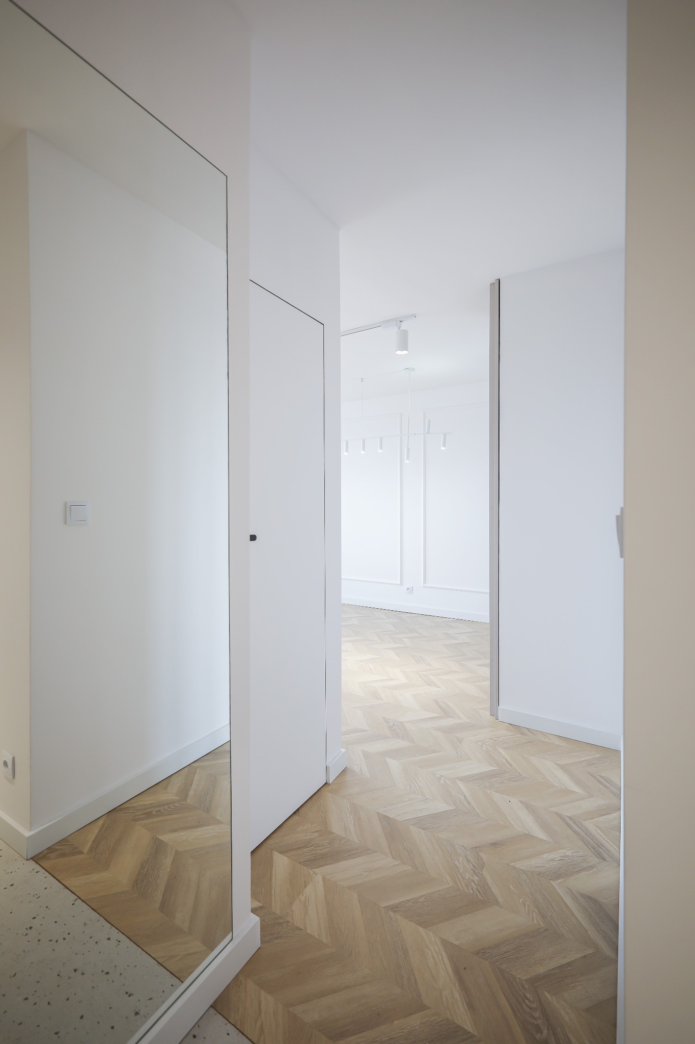 Wnętrze przedpokoju w minimalistycznym stylu z parkietem ułożonym we wzór jodełki, białymi ścianami i dużym lustrem.