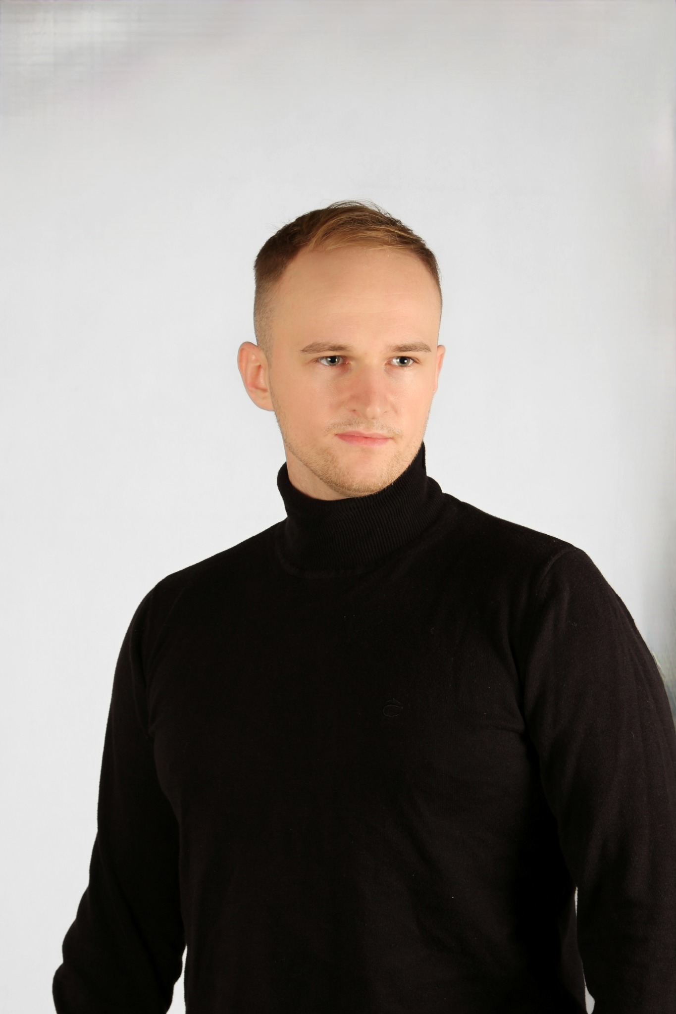 Portret młodego mężczyzny w czarnym golfie na jasnym tle, z delikatnym uśmiechem.