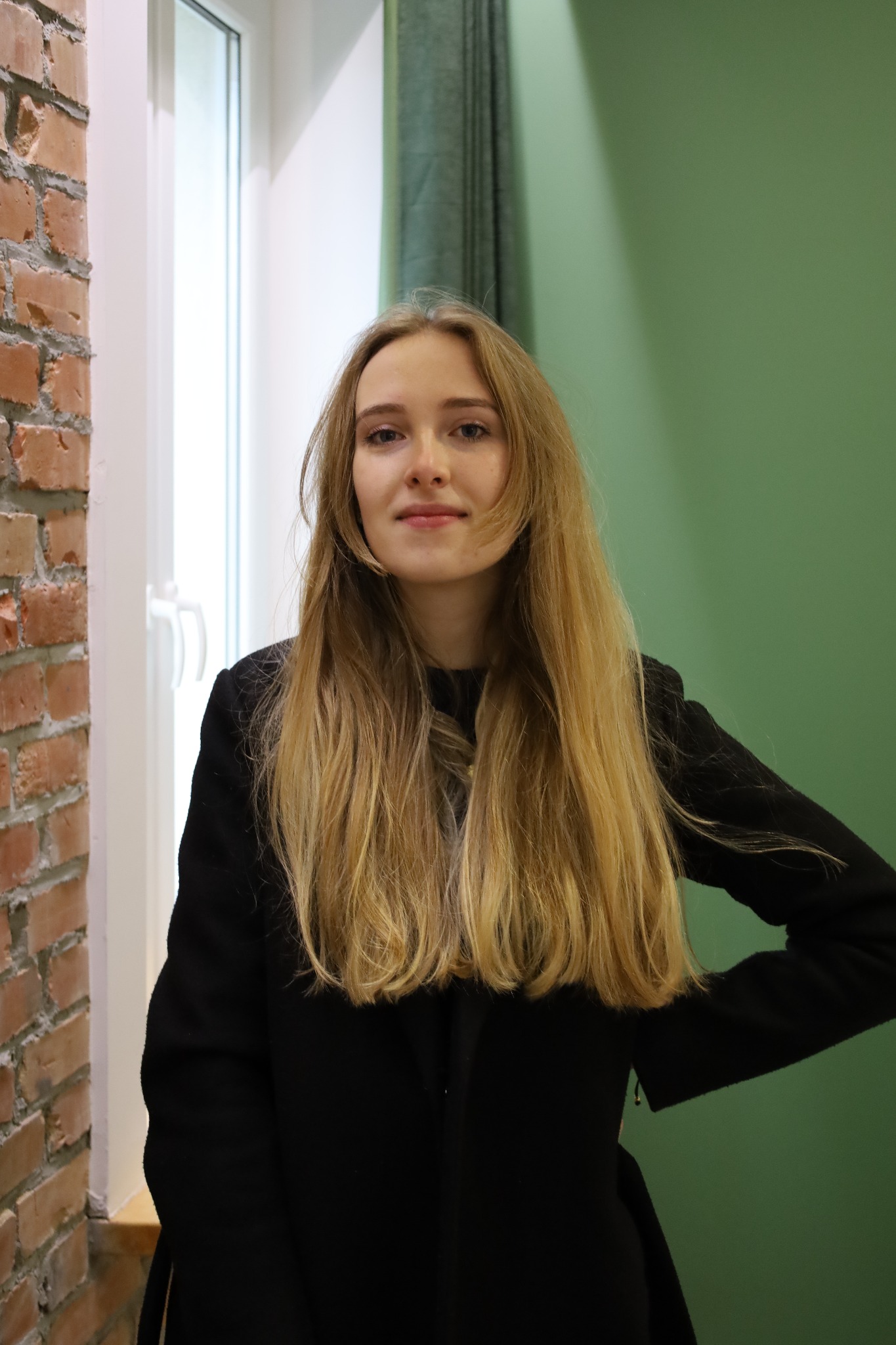 Portret młodej kobiety z długimi blond włosami na tle ceglanej ściany i zielonej ściany z zasłoną, w czarnym płaszczu, patrzącej w obiektyw.