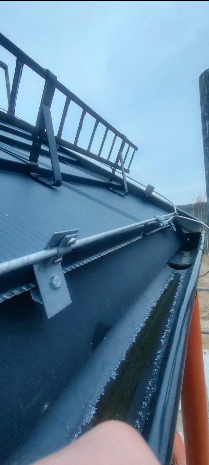 Zamocowany system odgromowy na dachu z blachy, widoczne uchwyty i przewody. Ujęcie z dołu, niebo w tle. Elementy metalowe w kolorze szarym.