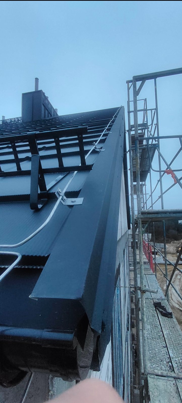 Czarny dach z zamontowaną instalacją odgromową, widoczne metalowe uchwyty i przewód. Ujęcie z dołu, w tle rusztowanie i budynek.