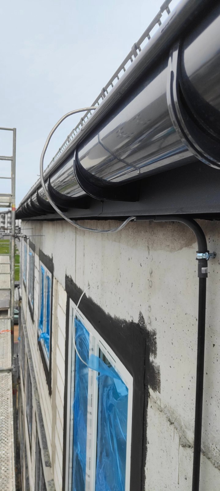 Montaż instalacji odgromowej na budynku w trakcie budowy. Widoczne rynny, przewody i okna zabezpieczone folią. Ujęcie z dołu, z widocznym fragmentem rusztowania.