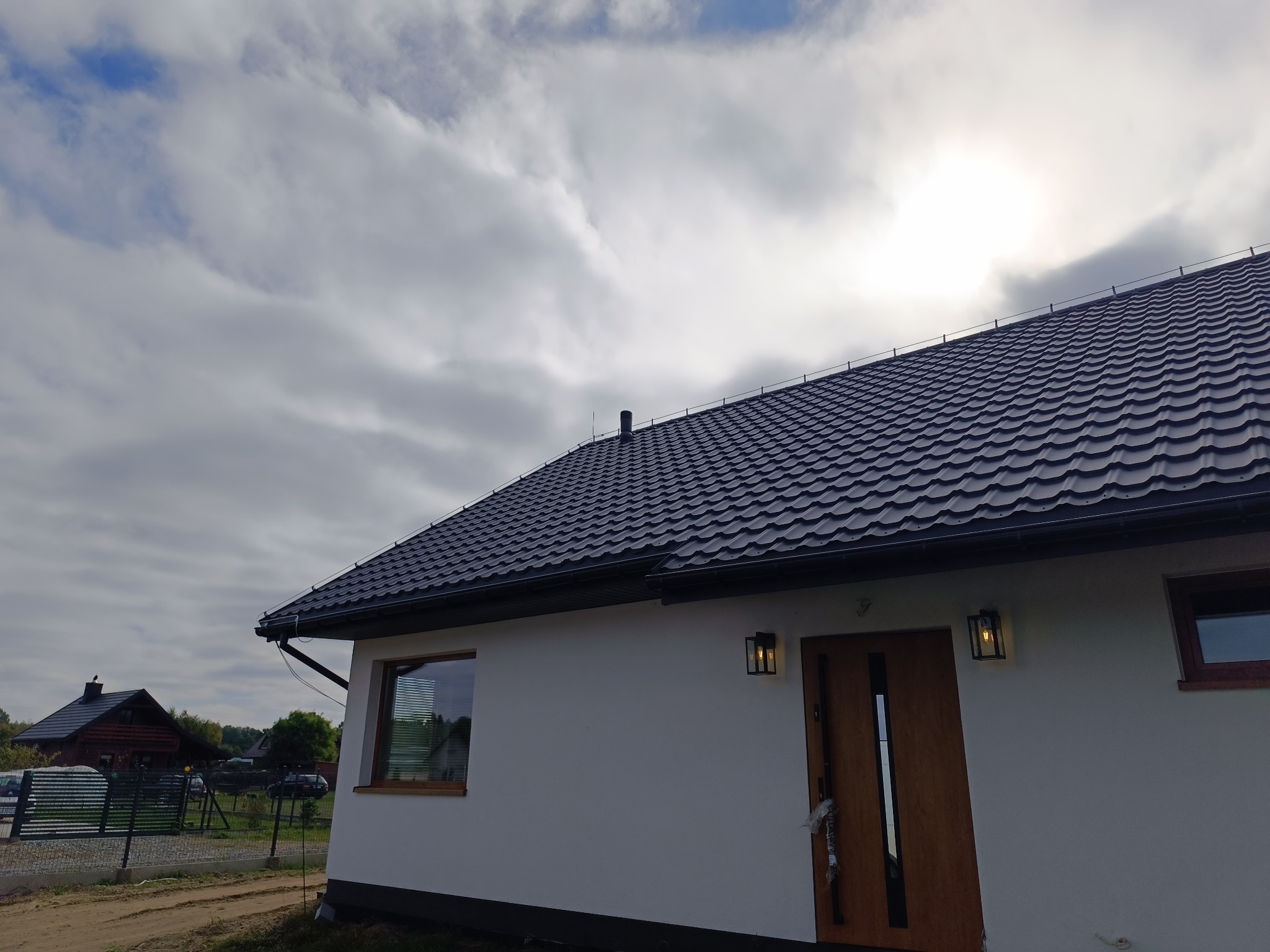 Nowoczesny dom jednorodzinny z ciemną dachówką i oświetleniem zewnętrznym. Widoczne okno z żaluzjami i drewniane drzwi wejściowe. W tle pochmurne niebo i fragment sąsiedniego budynku.