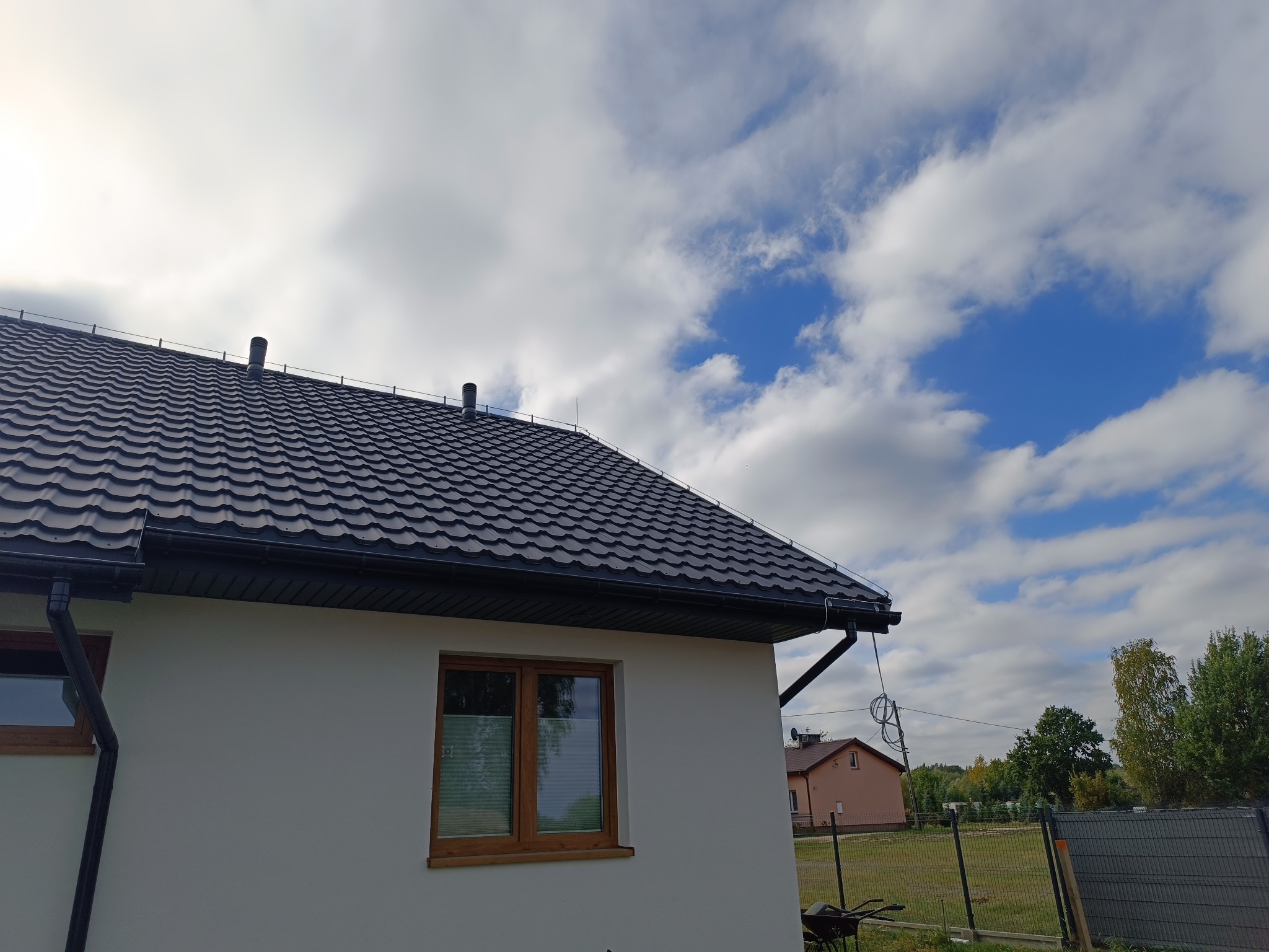 Nowy, grafitowy dach z orynnowaniem na jasnym domu jednorodzinnym, widok z dołu na tle błękitnego nieba z chmurami. Widoczne kominy wentylacyjne.