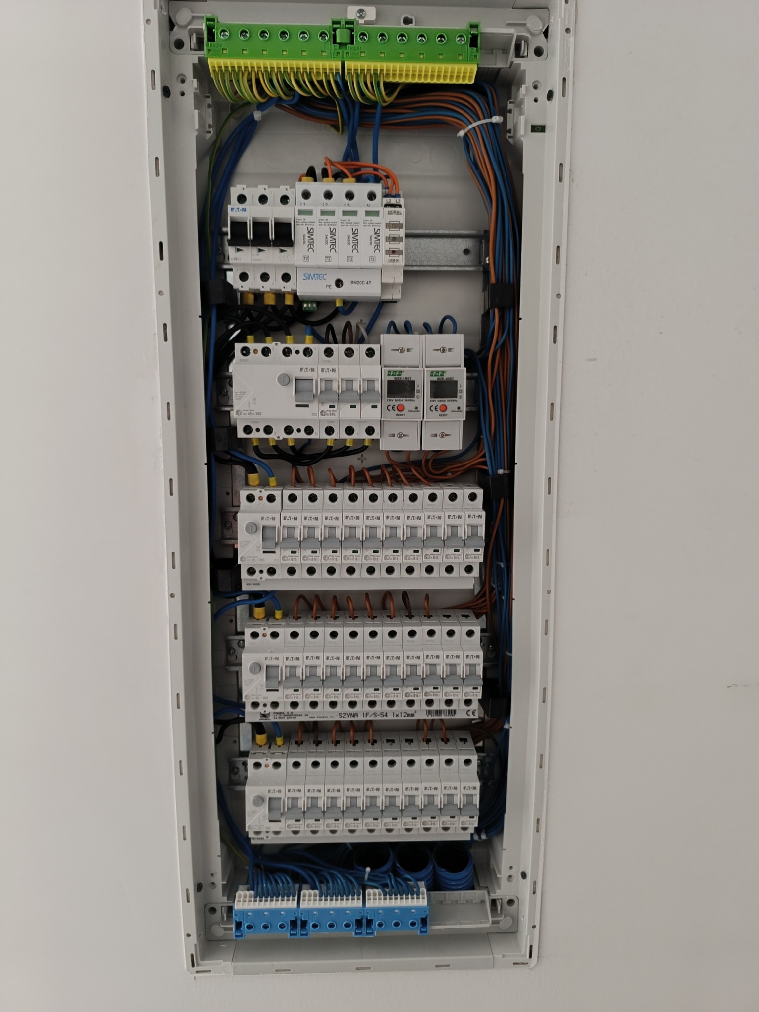 Rozdzielnica elektryczna w białej obudowie, widoczne zabezpieczenia, przewody w różnych kolorach, listwy zaciskowe. Instalacja schludna i uporządkowana.