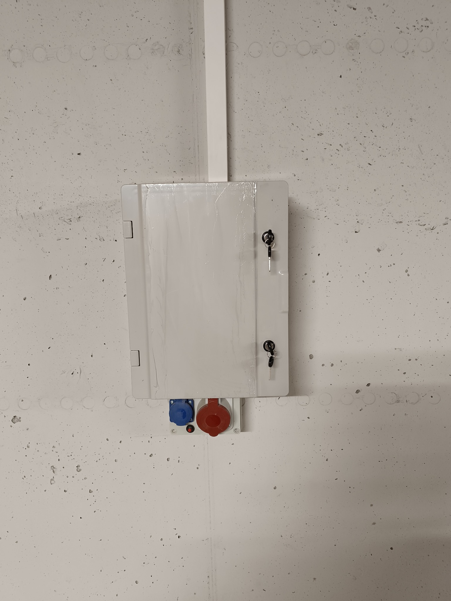 Biała skrzynka elektryczna z gniazdami (czerwone i niebieskie) zamontowana na szarej, betonowej ścianie. Widoczne klucze w zamkach. U góry biała listwa.