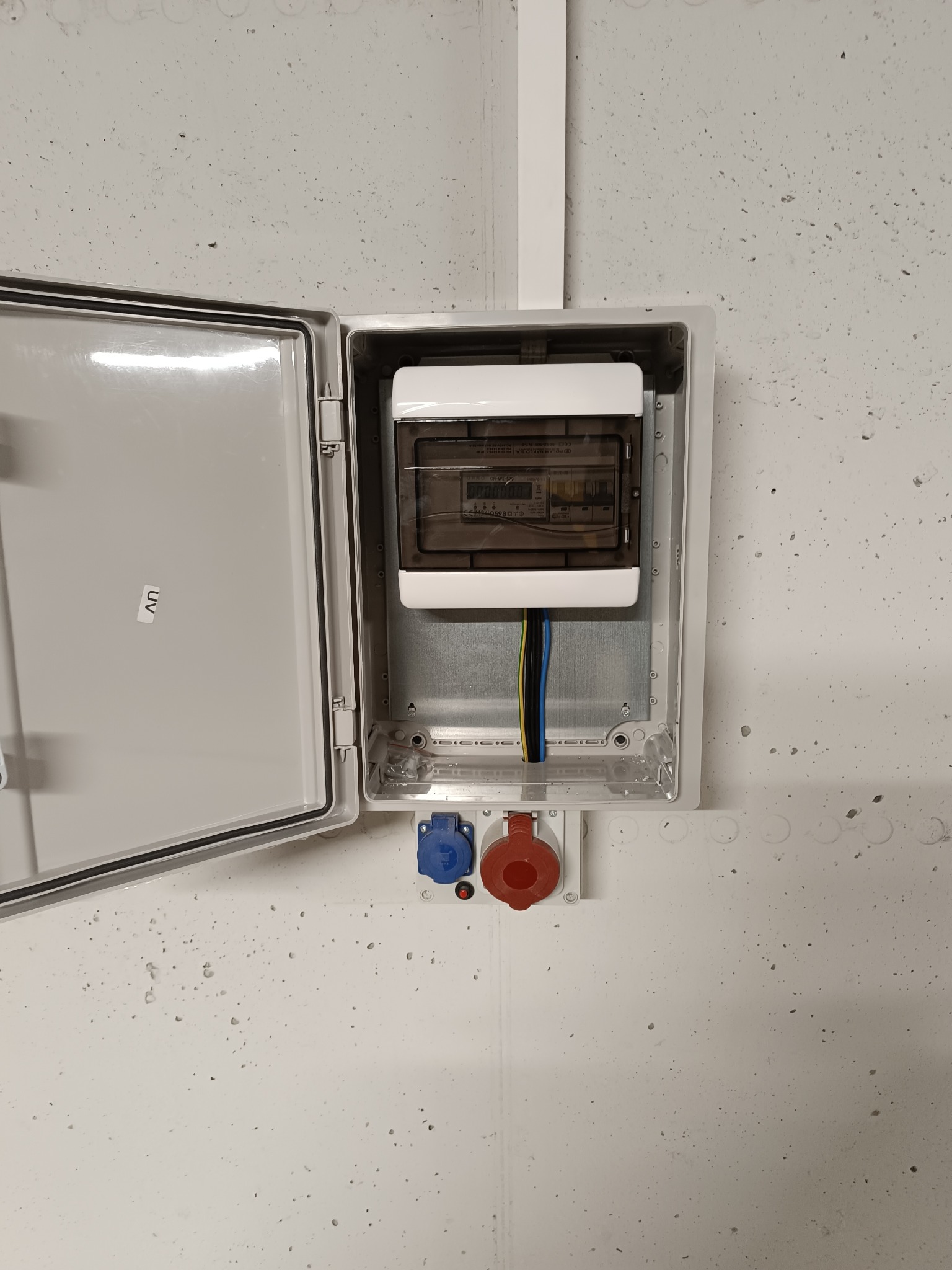 Otwarta skrzynka elektryczna z bezpiecznikami i gniazdami siłowymi na szarej, chropowatej ścianie. Widoczne przewody i elementy instalacji.