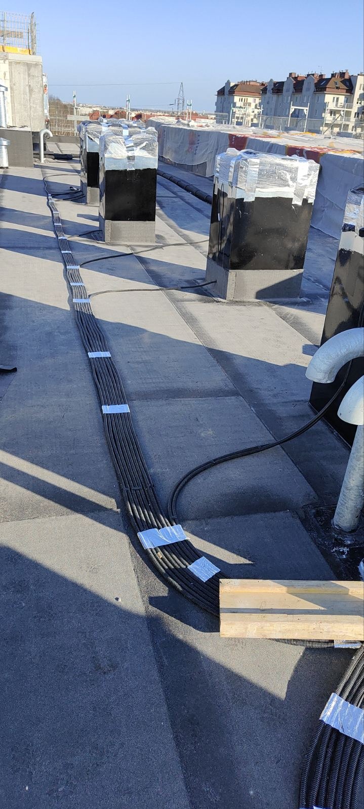 Układanie wiązki kabli w peszlach na dachu budynku, zabezpieczonych taśmą. W tle widoczne kominy wentylacyjne i budynki mieszkalne.