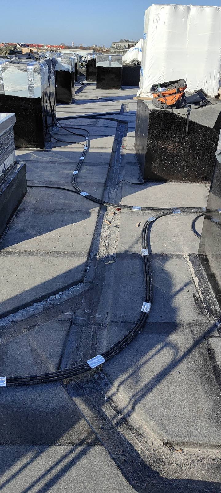 Instalacja kabli w peszlu na dachu budynku, zabezpieczone taśmą. Widoczne elementy wentylacji i narzędzia. Ujęcie z góry, w słoneczny dzień.