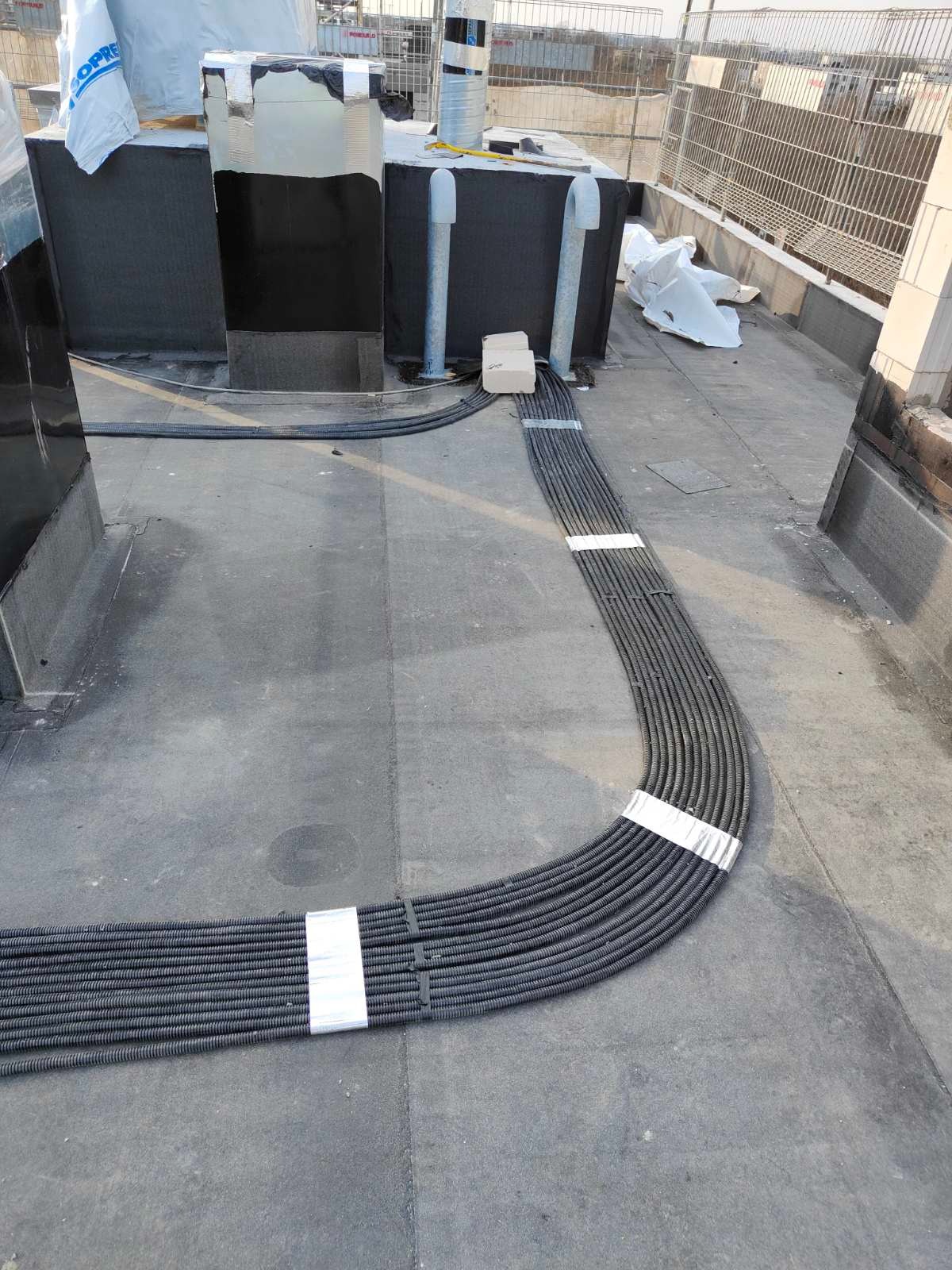 Równolegle ułożone, czarne rury kablowe na dachu, spięte taśmą. Widoczne elementy instalacji i fragmenty konstrukcji dachu. Ujęcie z góry, w dziennym świetle.