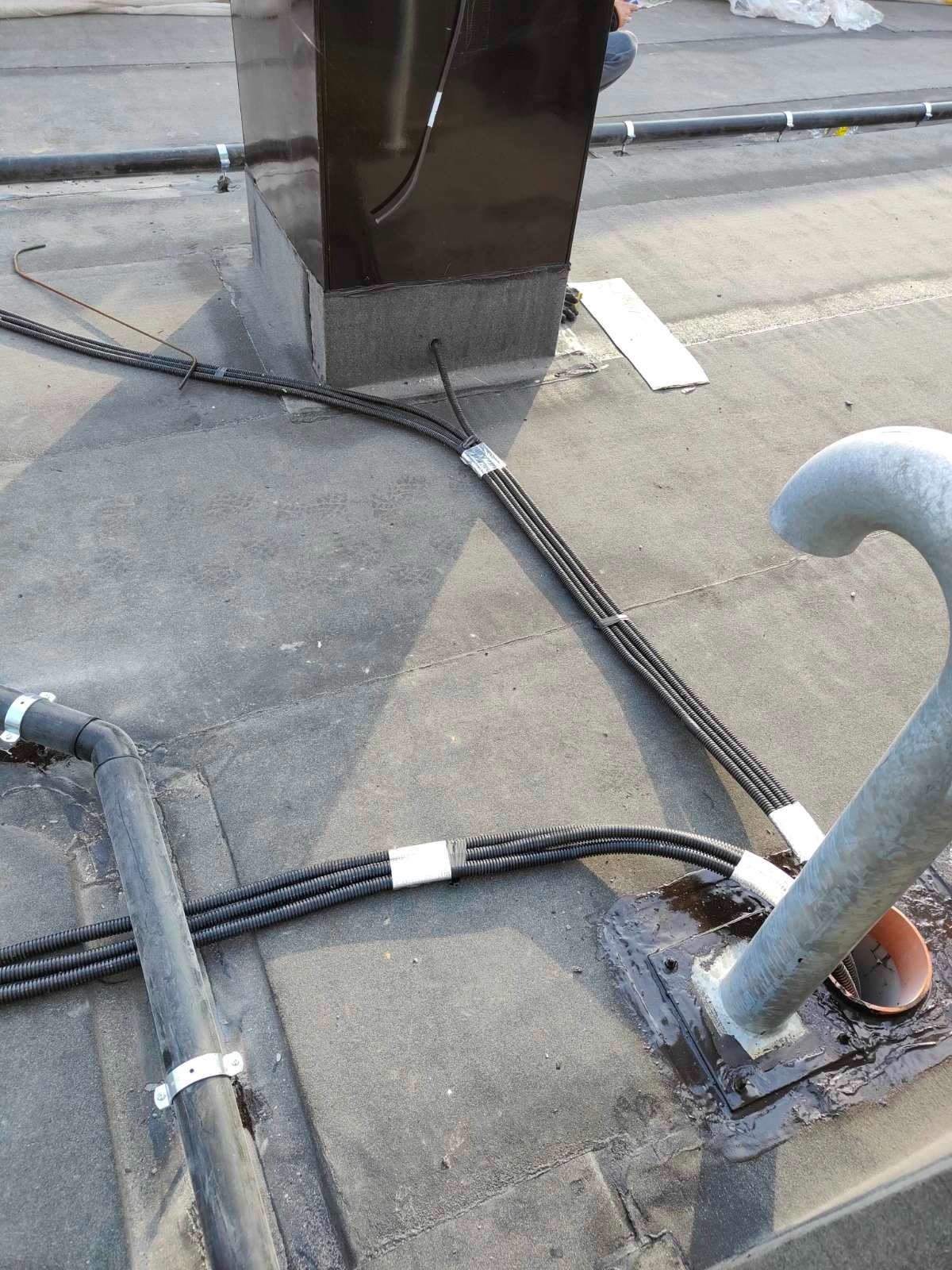 Instalacja elektryczna na dachu budynku z czarnymi rurami karbowanymi, zabezpieczonymi taśmą. Widoczny komin wentylacyjny i metalowy element instalacji odgromowej.