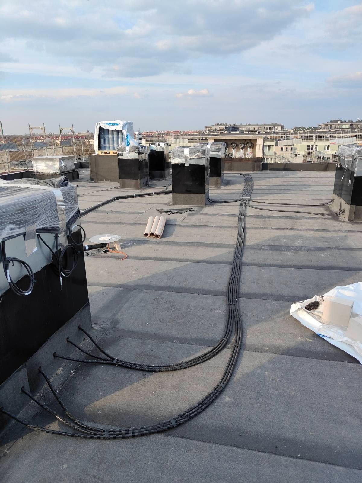 Instalacja przewodów w czarnej karbowanej osłonie na dachu budynku. Widoczne elementy wentylacyjne i inne instalacje. Przewody prowadzone równolegle, zabezpieczone uchwytami.