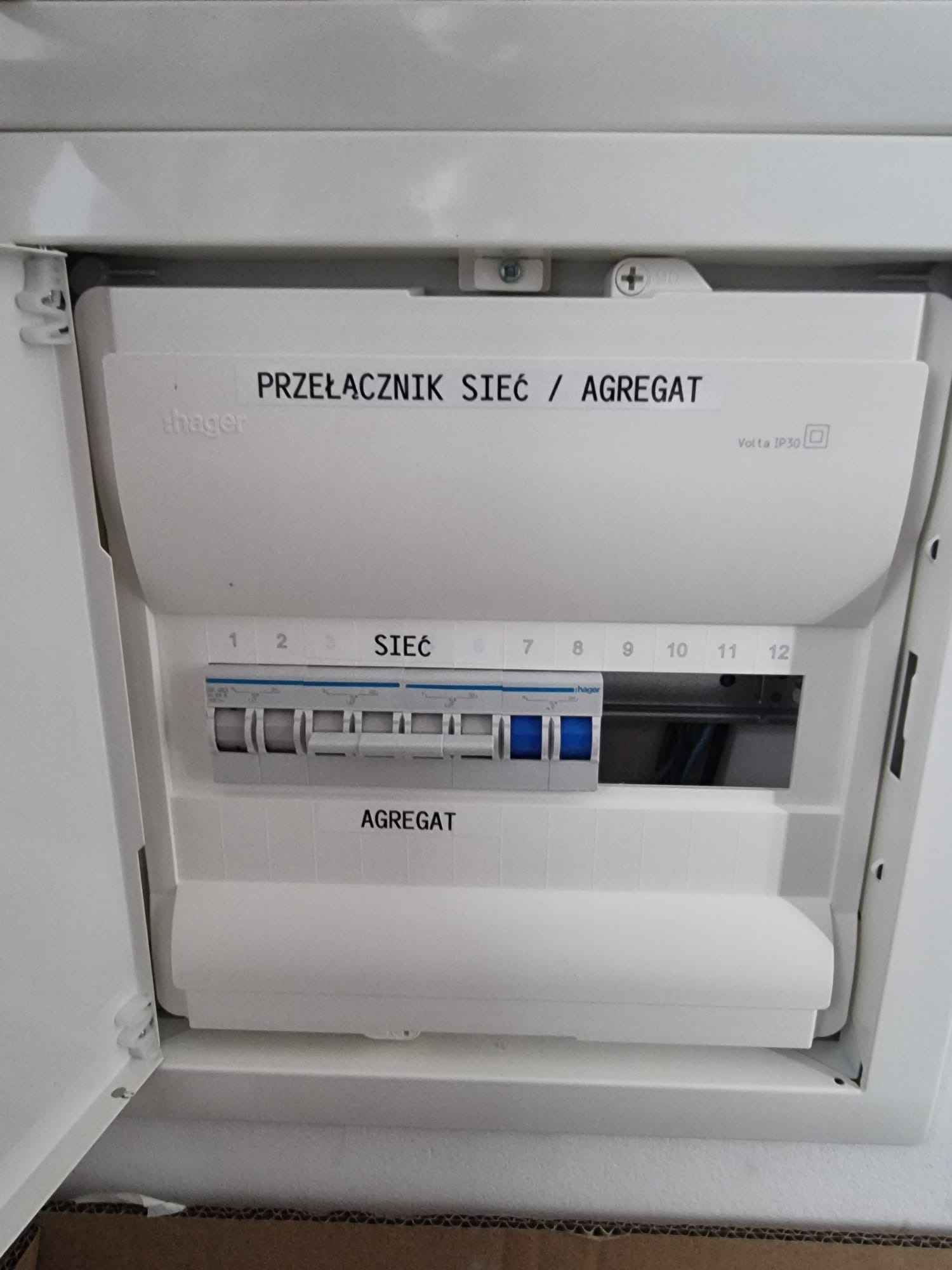 Biała skrzynka elektryczna Hager z przełącznikiem sieć/agregat, widoczne bezpieczniki i oznaczenia numerów. Otwarta obudowa ukazuje wnętrze.