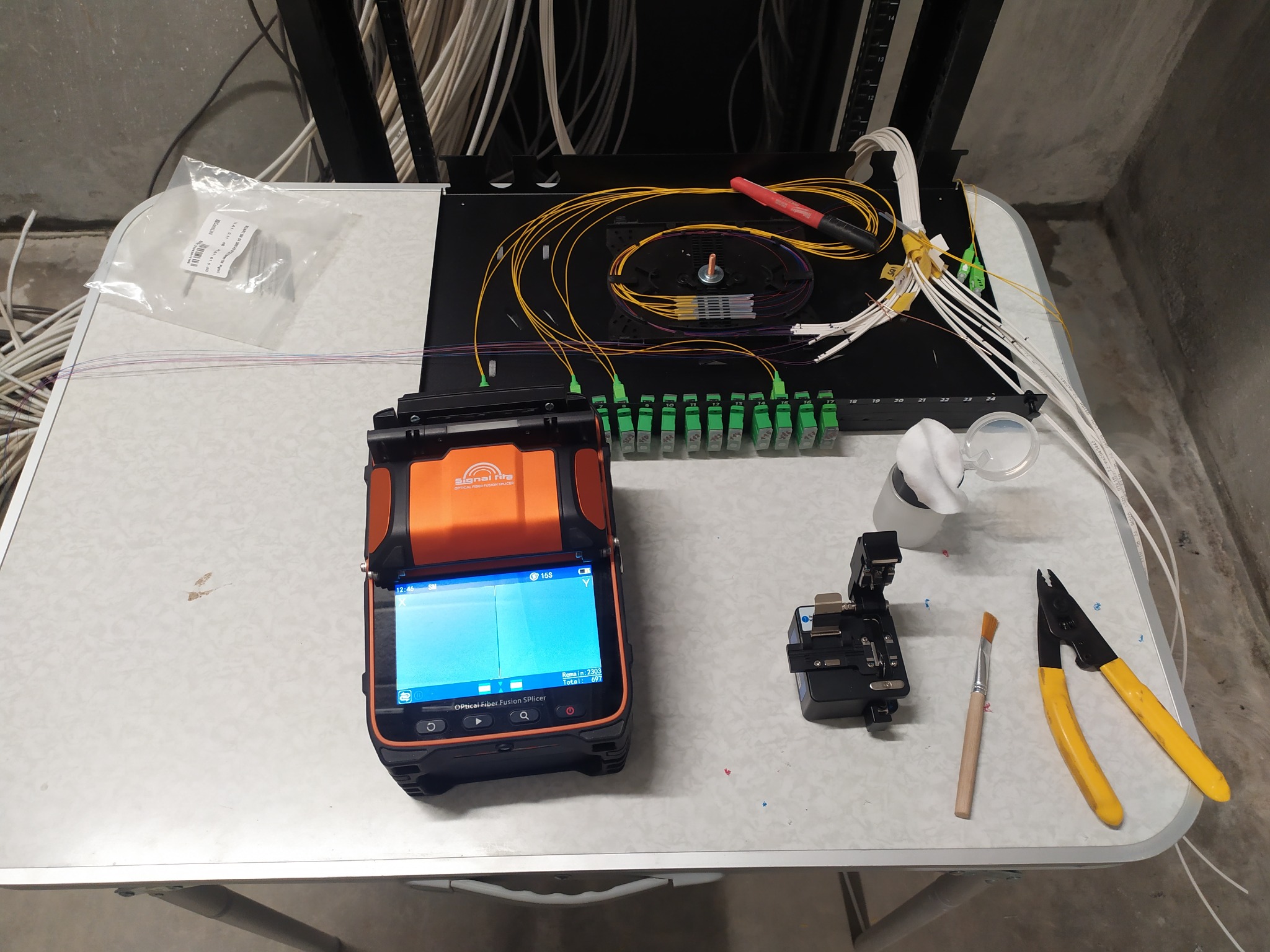 Sprzęt i narzędzia do spawania światłowodów na stole: spawarka światłowodowa, złączki, kable światłowodowe, obcinak, pędzelek. Praca w toku, precyzyjne łączenie.