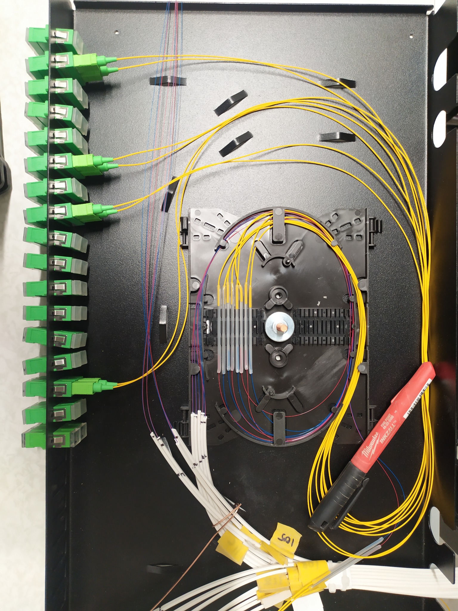 Rozdzielnica światłowodowa z zielonymi złączkami, żółtymi kablami i czarną tacką spawów. Widoczny czerwony marker. Precyzyjne połączenia światłowodowe.