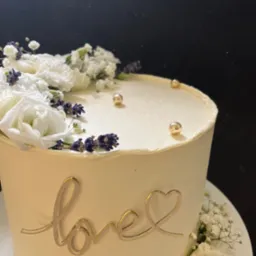 Elegancki tort z białym kremem, dekorowany białymi kwiatami, lawendą i złotymi kulkami, z napisem 'love' w złotym kolorze, na czarnym tle.