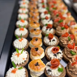 Rząd różnorodnych kanapek na ciemnej tacy: z kremowym serem i kawiorem, z pastą i pomidorem, oraz z ciasta francuskiego z czarnuszką.