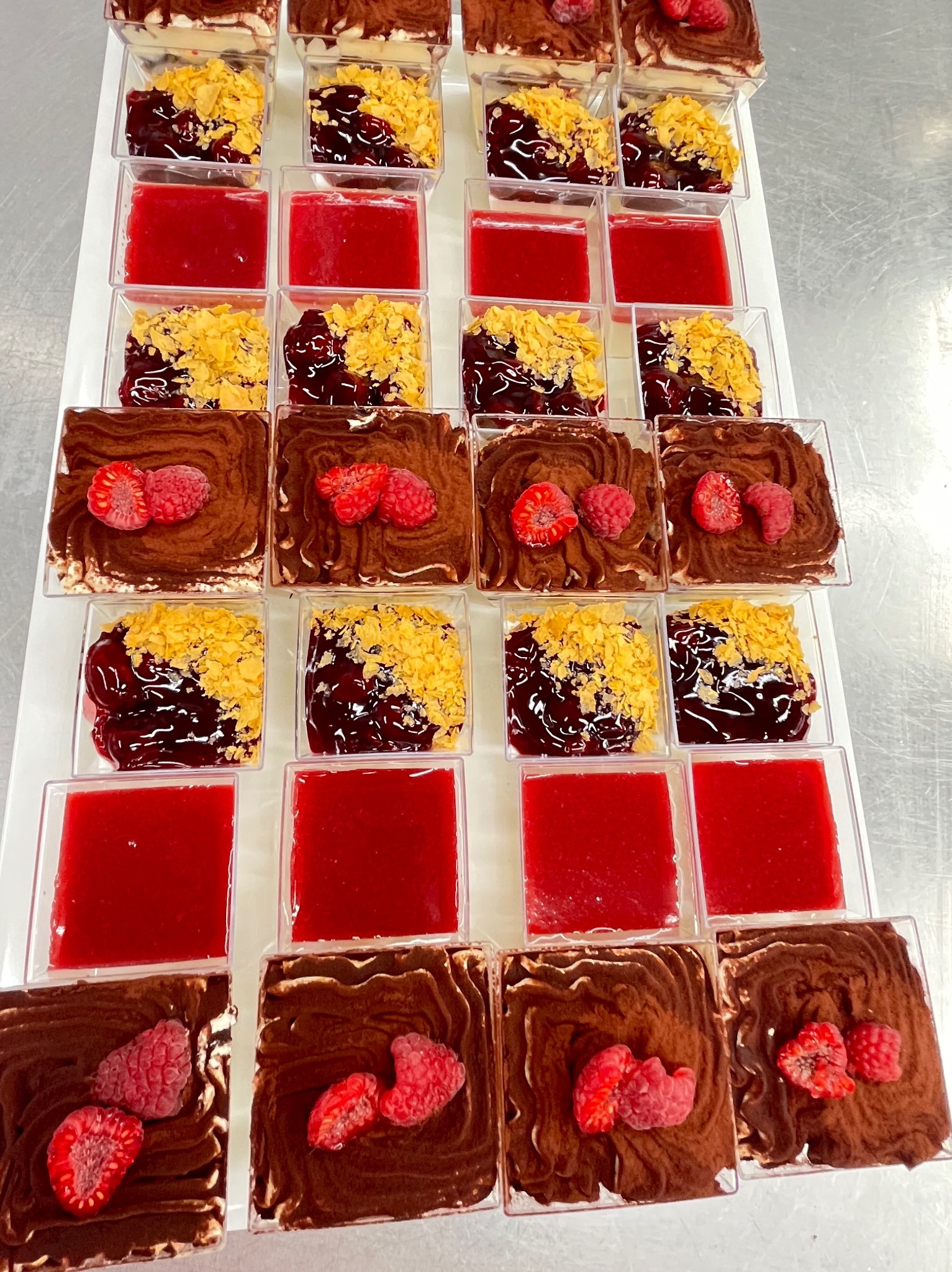 Rządki deserów w plastikowych pojemnikach: mus czekoladowy z malinami, galaretka wiśniowa z kruszonką i czysta galaretka truskawkowa na białej tacy.