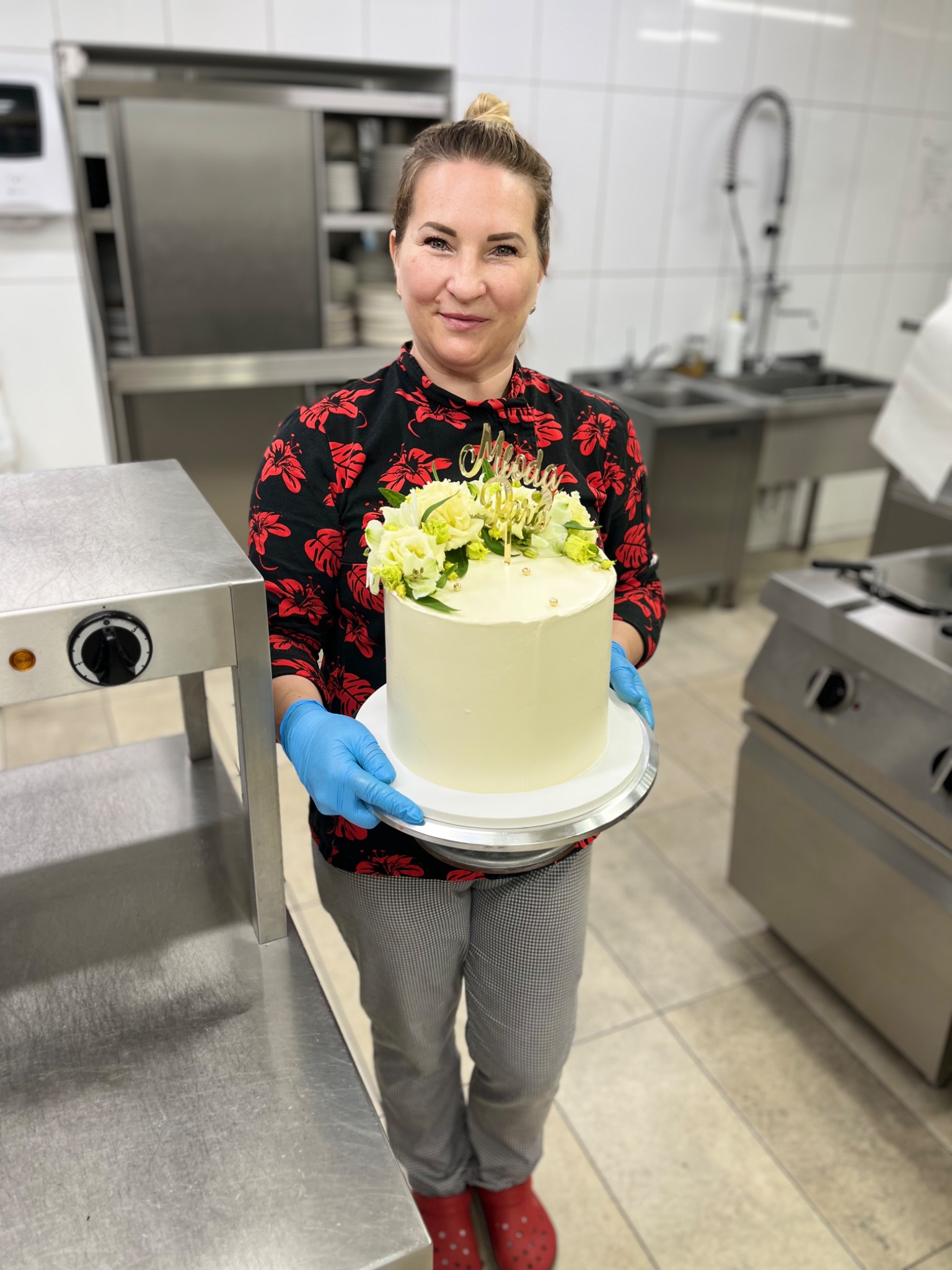 Uśmiechnięta kobieta w niebieskich rękawiczkach trzyma tort weselny z białym kremem, dekorowany świeżymi kwiatami i złotym napisem 'Młoda Para', w tle profesjonalna kuchnia.