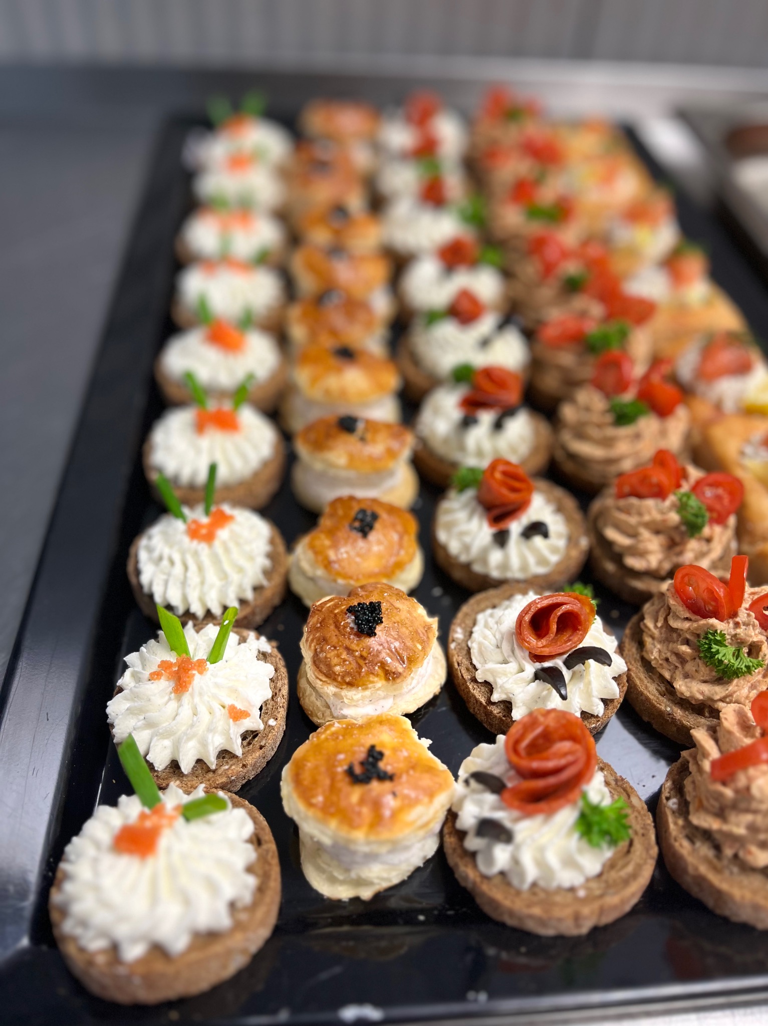 Rząd różnorodnych kanapek na ciemnej tacy: z kremowym serem i kawiorem, z pastą i pomidorem, oraz z ciasta francuskiego z czarnuszką.